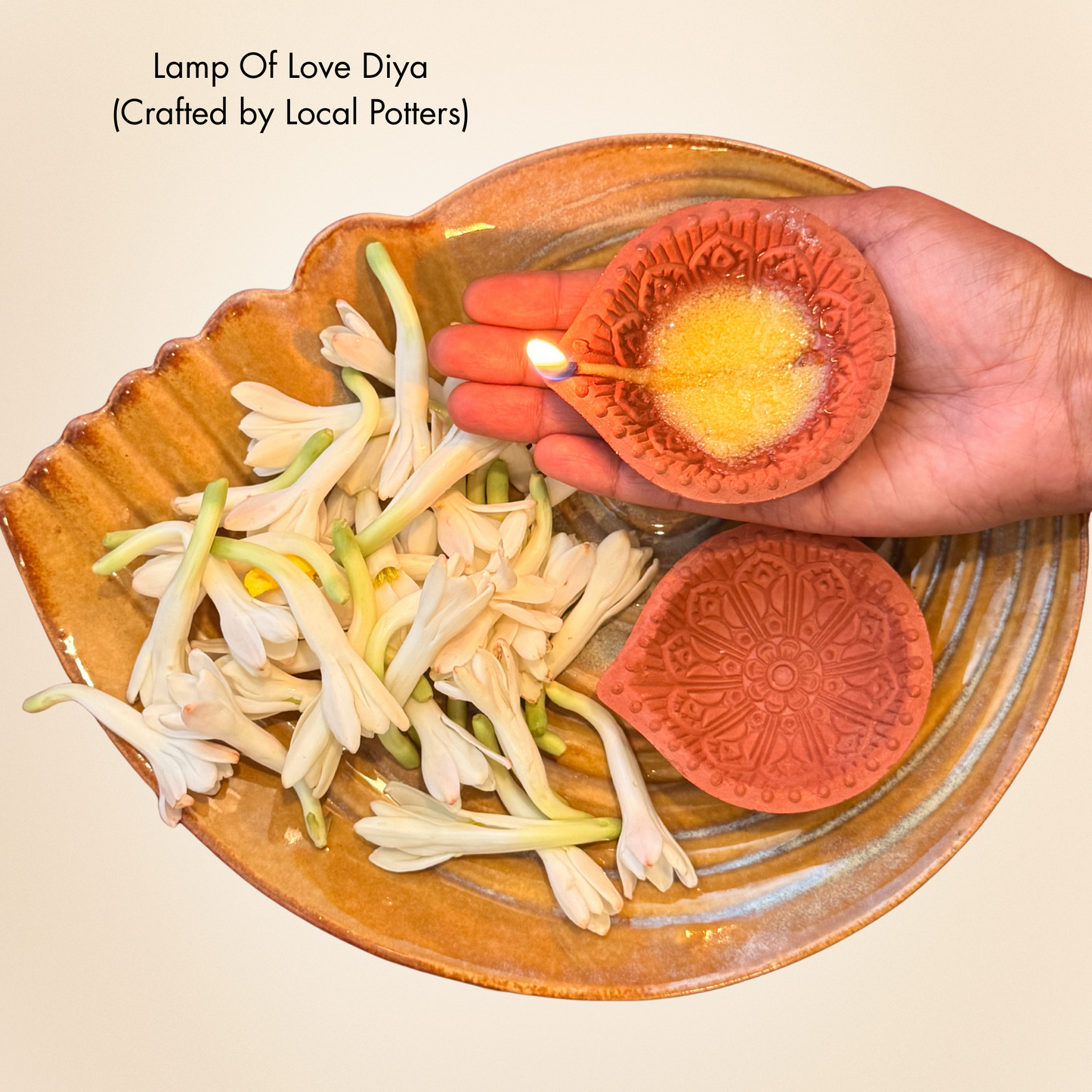 Ceramic diya being lit with a hand, surrounded by flowers on a wooden plate.