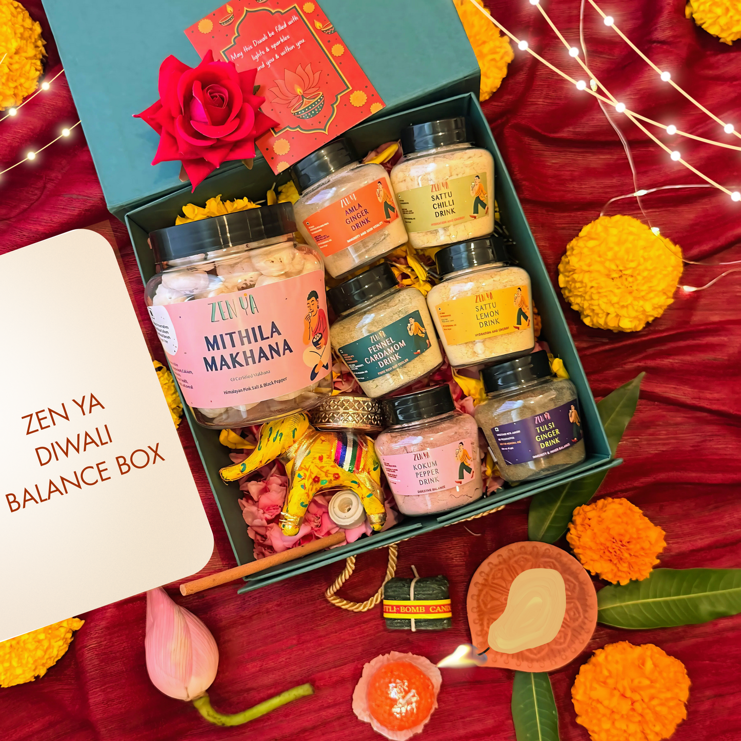 Zenya diwali wellness gift box. Mithila Makhana snack, zenya drinks, diya, candlesset in a decorative box with Diwali-themed elements on a red fabric background.