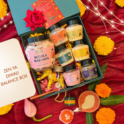 Zenya diwali wellness gift box. Mithila Makhana snack, zenya drinks, diya, candlesset in a decorative box with Diwali-themed elements on a red fabric background.